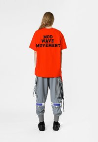 Camiseta roja de gran tamaño con "MOVIMIENTO MOD WAVE" impreso en negro en la parte posterior, combinada con pantalones cargo grises con múltiples bolsillos y correas.