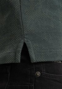 Mørkegrønt stof med et subtilt geometrisk mønster, der har sideslids ved kanten og er parret med sorte jeans og en metalknapdetalje.