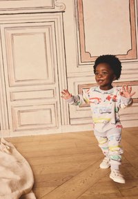 Hellgraues Baumwoll-Pyjama-Set mit bunten Autoaufdrucken. Besteht aus langärmligem Oberteil und Hose mit gerippten Bündchen. Kind steht auf Holzboden.