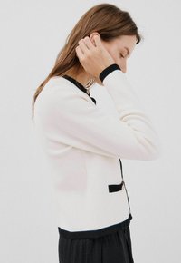 Cardigan beige lavorato a maglia con rifiniture nere, collo rotondo e dettagli con bottoni dorati. Design aderente, maniche lunghe e trama morbida.