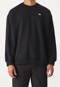 Schwarzer Baumwollpullover mit geripptem Rundhalsausschnitt, überschnittenen Schultern und einem weißen Logo-Akzent auf der Brust. Weicher, strukturierter Stoff.