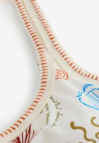 Detailný záber látky s viacfarebným potlačeným textom a vzormi, lemovaný krémovým a oranžovým tkaným okrajom.