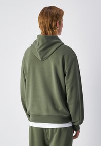 Sudadera color verde oliva de tejido suave, con una gran capucha, puños acanalados y un corte holgado. Lleva una camiseta blanca debajo.