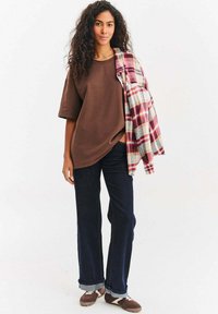 Brązowa koszula z krótkim rękawem, ciemnoniebieskie jeansy o szerokich nogawkach z podwiniętymi nogawkami oraz koszula flanelowa w kratę zarzucona na jedno ramię. Całość dopełniają brązowe trampki.