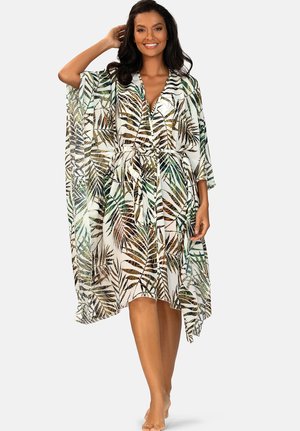 Uśmiechnięta kobieta w białej, przezroczystej sukience do kolan z zielonymi i brązowymi wzorami liści, pozująca z jedną ręką uniesioną do włosów.