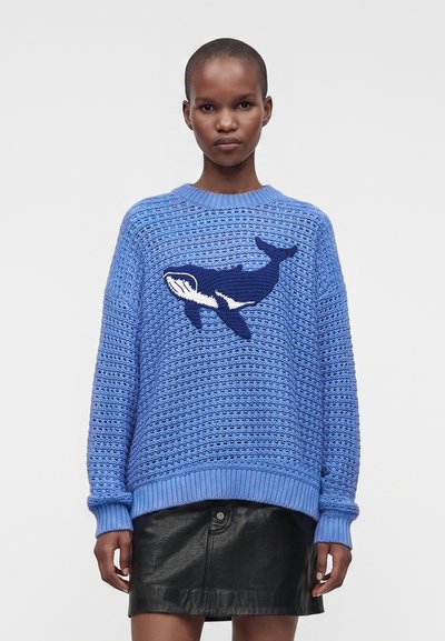 Personne portant un pull en tricot bleu avec un motif de baleine bleu foncé et une jupe en cuir noire, debout devant un fond clair uni.