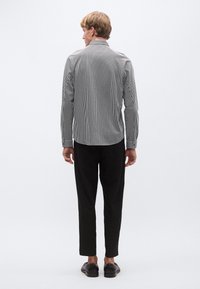 Melns un balts svītrains krekls ar garām piedurknēm, skats no aizmugures. Krekla apkakle ar pogām, gluda auduma, kombinēts ar melnām biksēm un kurpēm.