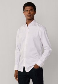 Mand iført en hvid langærmet skjorte med tynde lyserøde striber og mørke jeans, stående med den ene hånd i lommen.