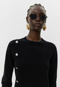 Granatowy sweter z dekoltem w szpic, bocznym detalem na guziki i miękką fakturą. Połączony z okrągłymi czarnymi okularami przeciwsłonecznymi oraz dużymi złotymi kolczykami.