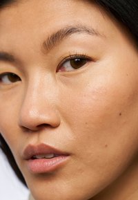 Gros plan sur un visage avec un teint lisse et uniforme. Les traits incluent des sourcils foncés, des yeux marron et une couleur de lèvres subtile avec de petites imperfections visibles.