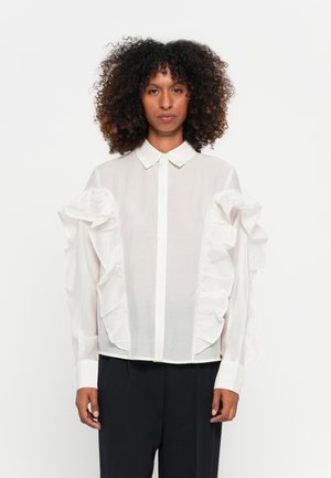 Camicia bianca con maniche arricciate, colletto e polsini aderenti, realizzata in tessuto leggero. Indossata con pantaloni scuri.