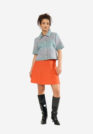 Žena oblečená v tyrkysovo vzorovanej krátkej bundovej blúzke na gombíky, oranžovej koleno dĺžkovej sukni a čiernych čižmách po kolená na bielom pozadí.