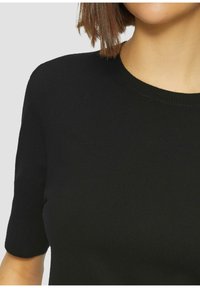 Frau trägt ein enganliegendes schwarzes kurzärmliges Strickoberteil mit Rundhalsausschnitt, gezeigt von den Schultern bis zur oberen Brust vor einem schlichten Hintergrund.