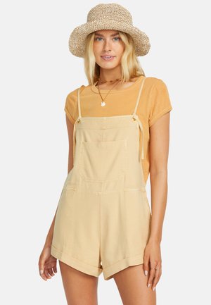 Blonde vrouw met een strooien hoed met brede rand, mosterdkleurig T-shirt met korte mouwen, beige mouwloze romper met korte broek en twee kettingen, staand tegen een witte achtergrond.