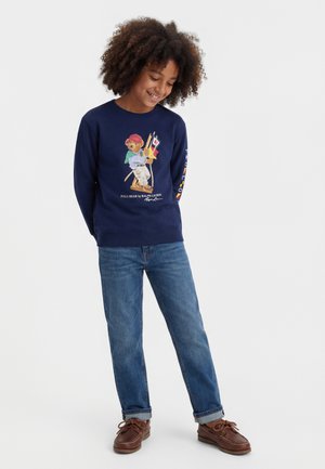 Otrok z kodrastimi lasmi, oblečen v mornarsko modro jopa z motivom medvedka, modre kavbojke in rjavi čevlji, se smehlja in gleda navzdol.