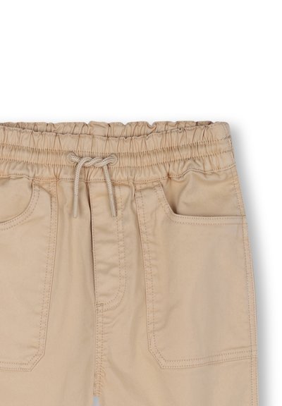 Pantalons beiges à taille élastique avec cordon de serrage, présentant deux poches avant et des détails de couture visibles. Texture de tissu en coton lisse.