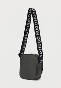 Συμπαγής μαύρη τσάντα χιαστί με φαρδύ μαύρο λουρί που εμφανίζει επαναλαμβανόμενα λευκά λογότυπα "HUGO", και κλείσιμο με φερμουάρ σε απλό φόντο.