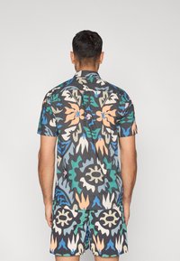 Πουκάμισο με κοντά μανίκια και σορτς από σκούρο ύφασμα με πολύχρωμο αφηρημένο λουλουδάτο μοτίβο σε πράσινους, μπλε, ροδακινί και λευκούς τόνους.