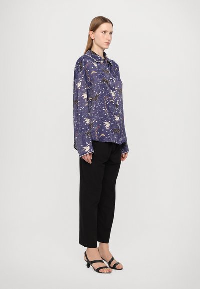 Cămașă cu mânecă lungă din material violet, cu modele cerești, închidere cu nasturi și manșete contrastante, asortată cu pantaloni negri largi și sandale.