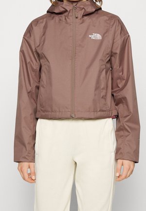 Braune North Face Kapuzenjacke mit Reißverschluss, getragen zu cremefarbenen Hosen, gezeigt von den Schultern bis zur Mitte des Oberschenkels, stehend vor einfarbigem Hintergrund.