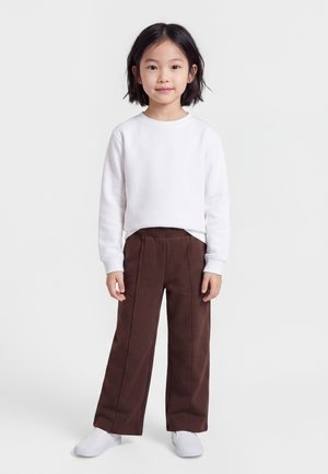 Jeune fille debout, portant un t-shirt blanc à manches longues, un pantalon marron large et des baskets blanches sur un fond clair uni.