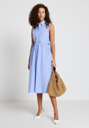 Femme aux cheveux bouclés portant une robe midi sans manches bleu clair à fines rayures, des talons blancs, et tenant un grand sac tressé beige.