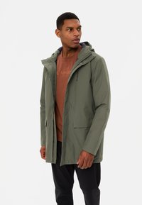Olivegroene waterdichte jas met een capuchon, ritssluiting, twee voorkeurszakken en een gladde textuur, gedragen over een bruine T-shirt.
