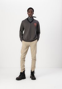 Grå sweatshirt med V-ringning och röd grafik, ihop med beige byxor och svarta ankelboots. Outfitten inkluderar en svart och vit mönstrad bandana.