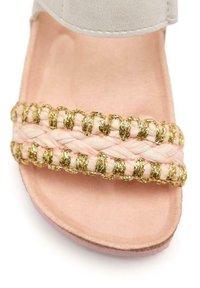 Sandalo rosa chiaro con tomaia in suede e una cinturino intrecciato con dettagli dorati. Suola morbida e piatta con una finitura opaca e liscia.