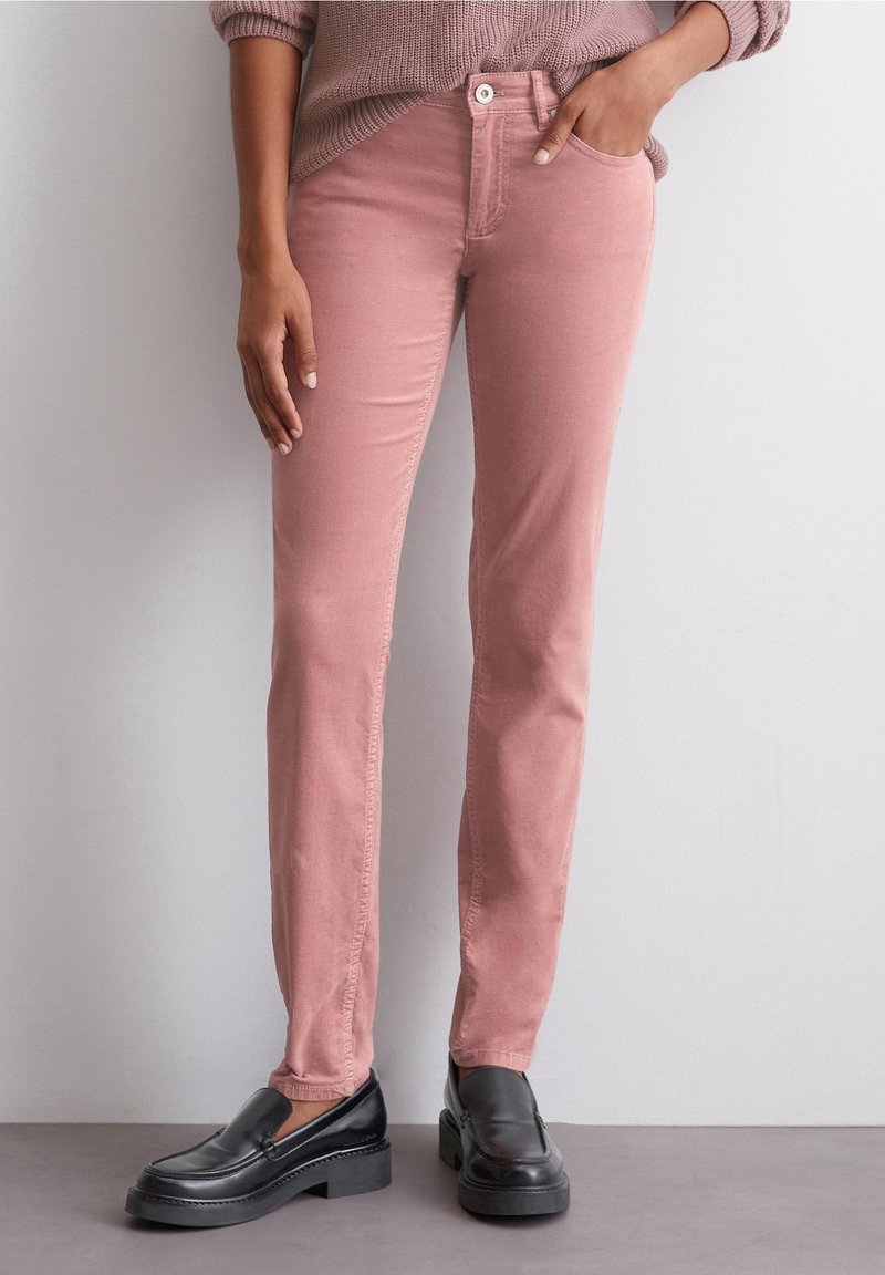Pantalon slim rose poudré porté avec un pull mauve et des mocassins en cuir noir, mains détendues le long du corps.