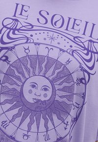 Camiseta de algodón morada con un gráfico de sol circular que tiene una cara, rodeado de símbolos zodiacales y espirales decorativas.