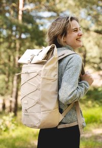 Sac à dos beige au design à rabat, en tissu texturé, avec des bretelles réglables et des détails en cordon élastique, porté par une personne dans une zone boisée.
