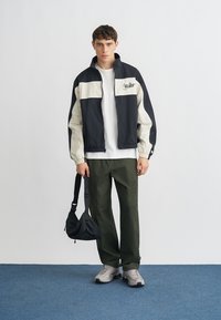 Μαύρο και κρεμ ζακέτο Nike με φερμουάρ και λογότυπο, συνδυασμένο με λευκό μπλουζάκι και χακί παντελόνι. Γκρι αθλητικά παπούτσια και μαύρη τσάντα ολοκληρώνουν την εμφάνιση.