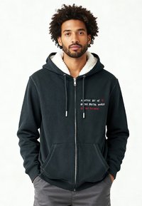 Hoodie zippé gris foncé avec une capuche doublée en polaire, deux poches avant et un texte brodé sur la poitrine dans des couleurs contrastantes.