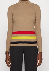 Jersey de cuello alto beige con canalé que presenta atrevidas rayas horizontales en rojo, amarillo y negro en la mitad inferior. Incluye un detalle de logo.