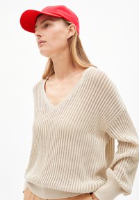 Beige knitted sweater with a V-neck and loose sleeves, featuring a ribbed texture, paired with a bright red cap.