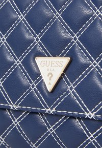 Mornarska prešita torbica iz sintetične usnja z belimi šivi in trikotno zlatim logotipom s napisom "GUESS".