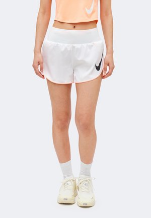 Kobieta ubrana w białe krótkie spodenki do biegania Nike, jasnopomarańczowy krótki top, białe skarpety za kostkę i kremowe buty sportowe Nike, stoi z rozluźnionymi rękami.