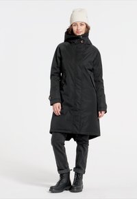 Zwarte, waterdichte jas tot de knie met een hoge kraag, ritssluiting aan de voorkant en zijzakken. Gecombineerd met donkerblauwe jeans en zwarte laarzen.