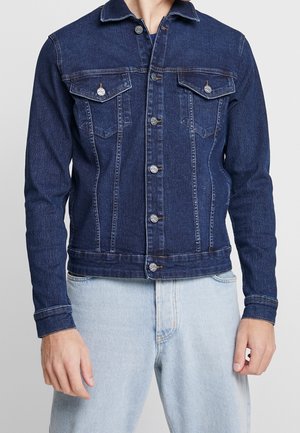 Donkerblauwe spijkerjack met knopen aan de voorkant en klepzakken op de borst, gedragen over lichtblauwe spijkerbroek door een staand persoon.