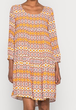 Frau trägt ein langärmliges, knielanges Kleid mit gelben und pinken Blumen- und geometrischen Mustern auf weißem Hintergrund.