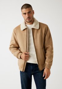 Beige Bomberjacke mit weißem Kunstfellkragen, Reißverschluss vorne, gerippten Bündchen und seitlichen Taschen, getragen über einem hellen Strickpullover und dunklen Jeans.