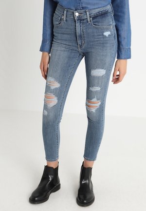 Slim-fit, light blue denim jeans featuring distressed detailing with rips, five-pocket design, and a high waist, paired with black ankle boots.
