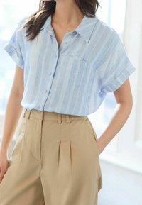 Chemise à manches courtes bleu clair avec boutons, rayures verticales blanches ; dotée d'une poche poitrine et d'une coupe décontractée, associée à un pantalon beige plissé.