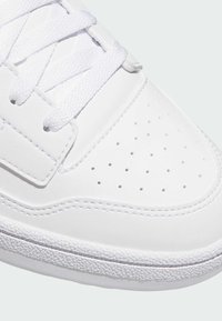 Biele športové topánky s hladkým syntetickým zvrškom, perforovanou špičkou, štruktúrovanou podrážkou a plochými bielymi šnúrkami. Jednoduchý, minimalistický dizajn.