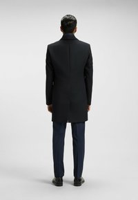 Cappotto nero con design sartoriale, spacco singolo sul retro, abbinato a pantaloni blu navy e scarpe nere, mostrato di spalle.