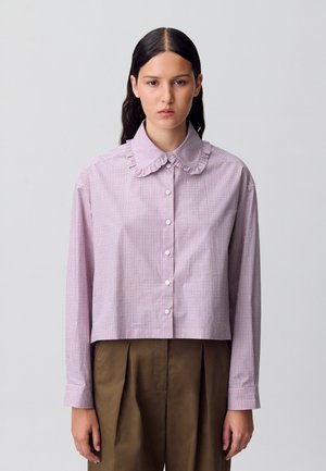 Chemise courte, à boutons, en vichy rose et blanc. Présente un col à volants et des manches longues. Tissu en coton texturé.
