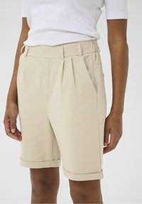 Shorts beige sur mesure avec une taille haute, des plis et des ourlets roulés. Texture de tissu lisse, longueur mi-cuisse, avec poches latérales.
