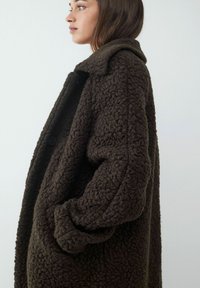 Bruine gestructureerde oversized jas met een grote kraag, knoopsluiting en grof stof, zijaanzicht getoond.