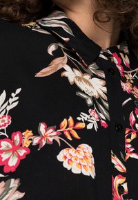 Zbliżenie czarnej koszuli z guzikami w multikolorowe wzory kwiatowe i kołnierzykiem, noszonej przez osobę z kręconymi włosami.